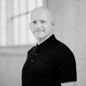 Black-and-white portrait of a bald man in a dark polo shirt standing indoors.